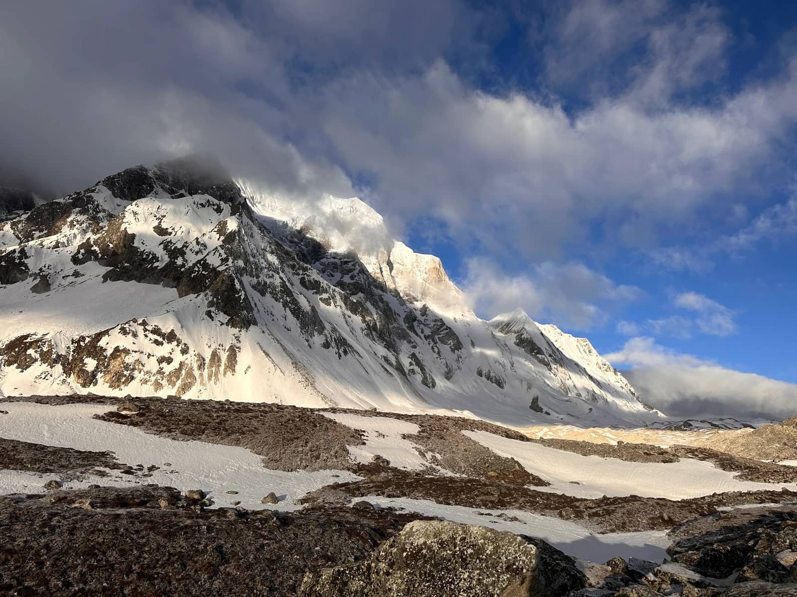 A rugged, snow-covered mountain ridge is partially obscured by swirling clouds against a blue sky.
