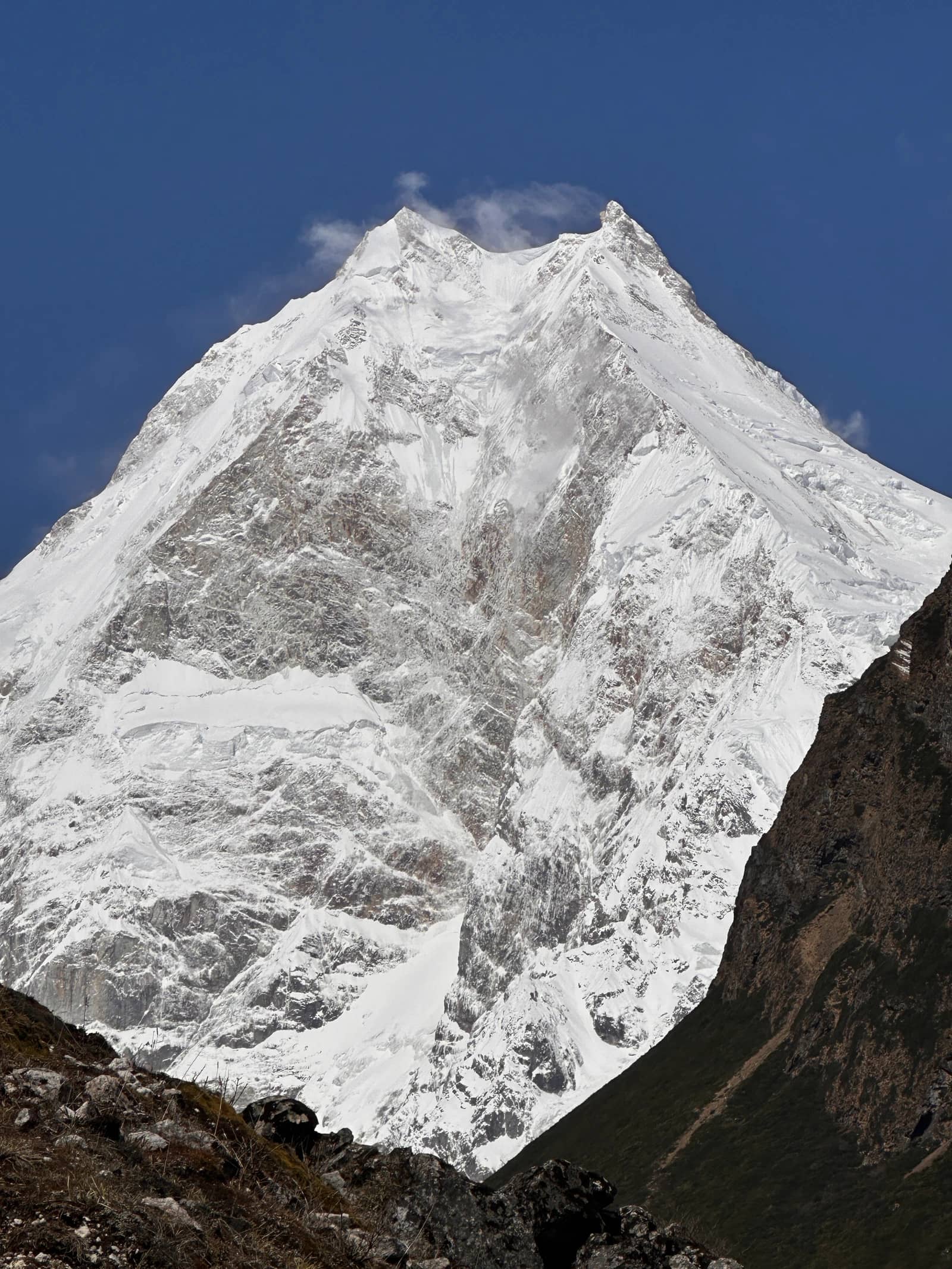 A towering, snow-covered mountain peak dominates a deep blue sky, framed by dark foreground slopes.