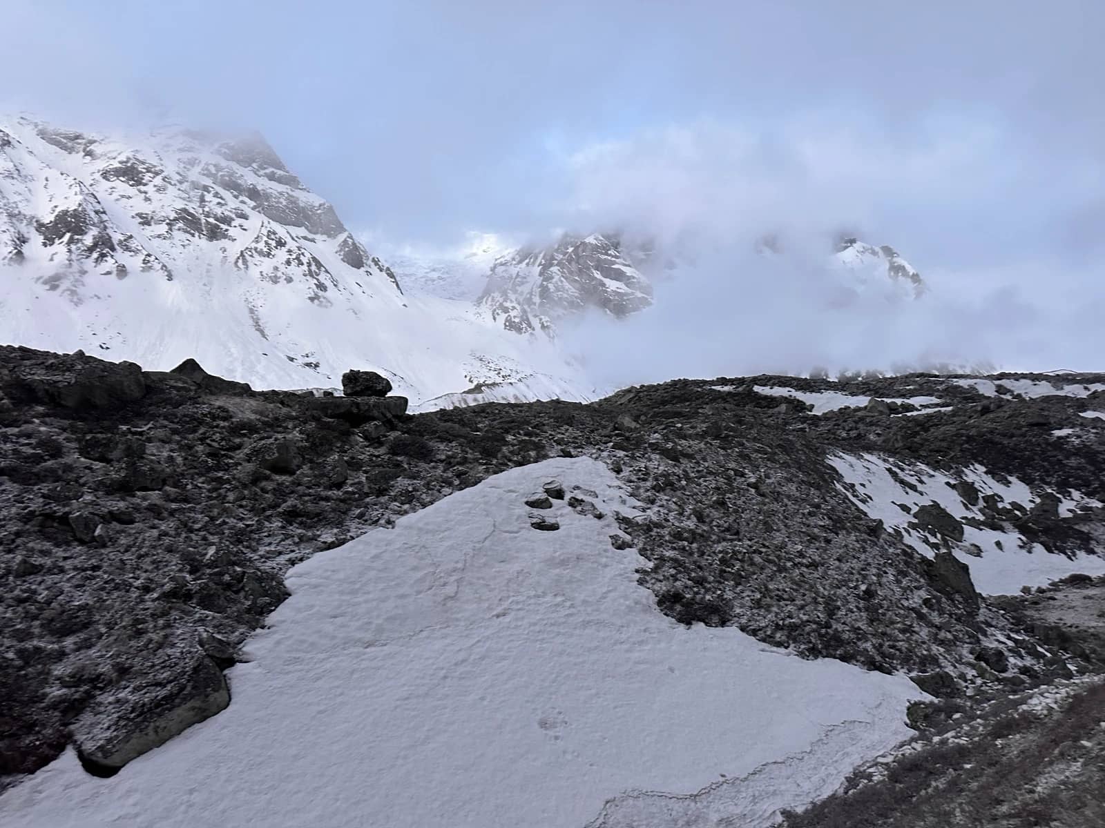 A rocky, snow-covered landscape with a faint trail leading toward mist-shrouded mountain peaks.