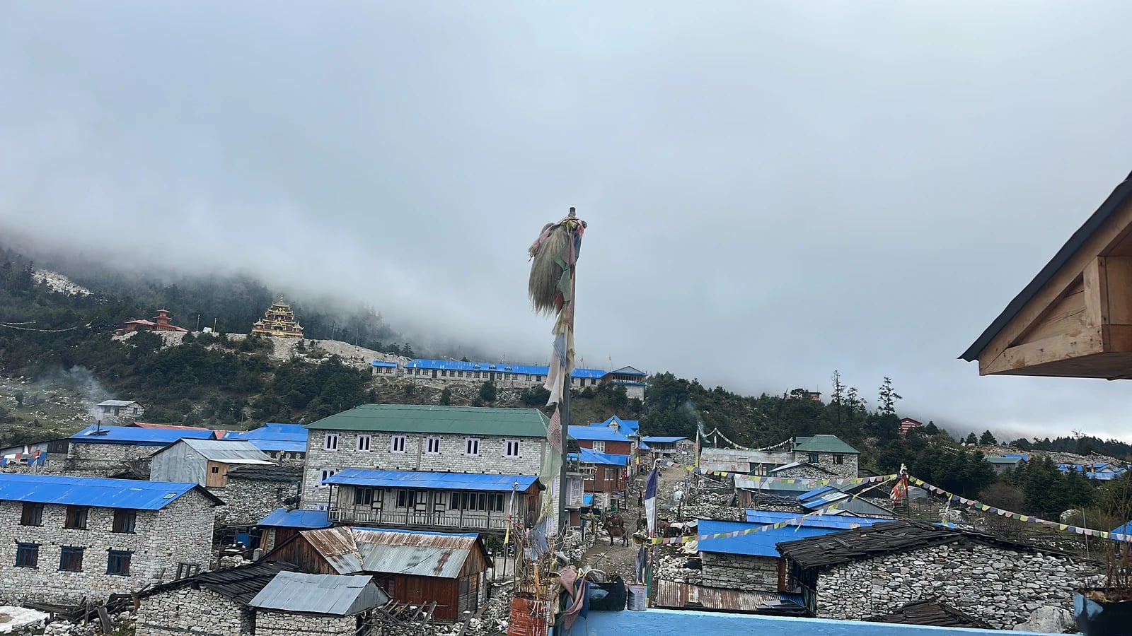 A stone village with blue roofs sits beneath a misty hillside featuring a golden monastery and tall prayer flag pole.