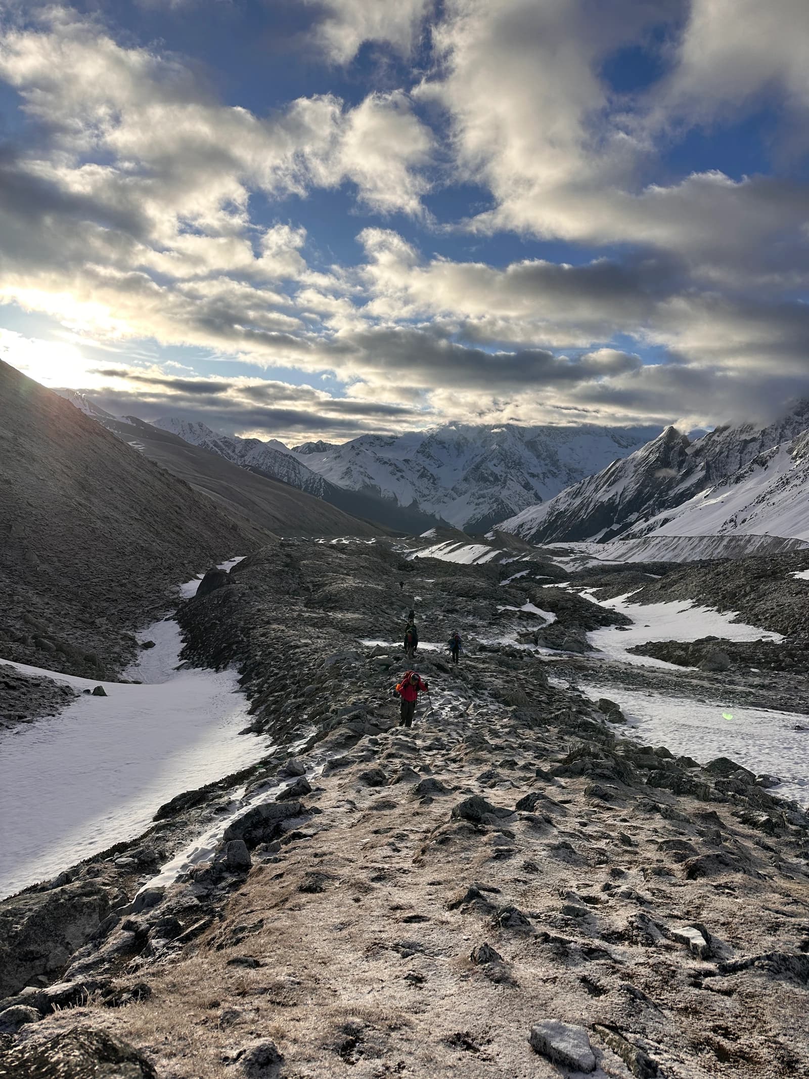 Trekkers hike along a rocky ridge through a snowy mountain landscape under a dramatic cloudy sky.