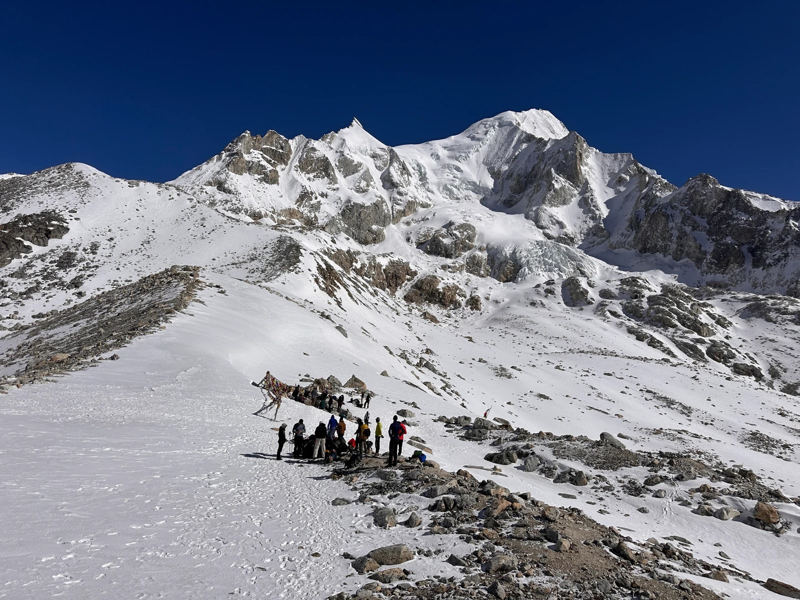 A group of trekkers gathers on a snowy slope beneath a towering, rugged mountain peak under a clear blue sky.