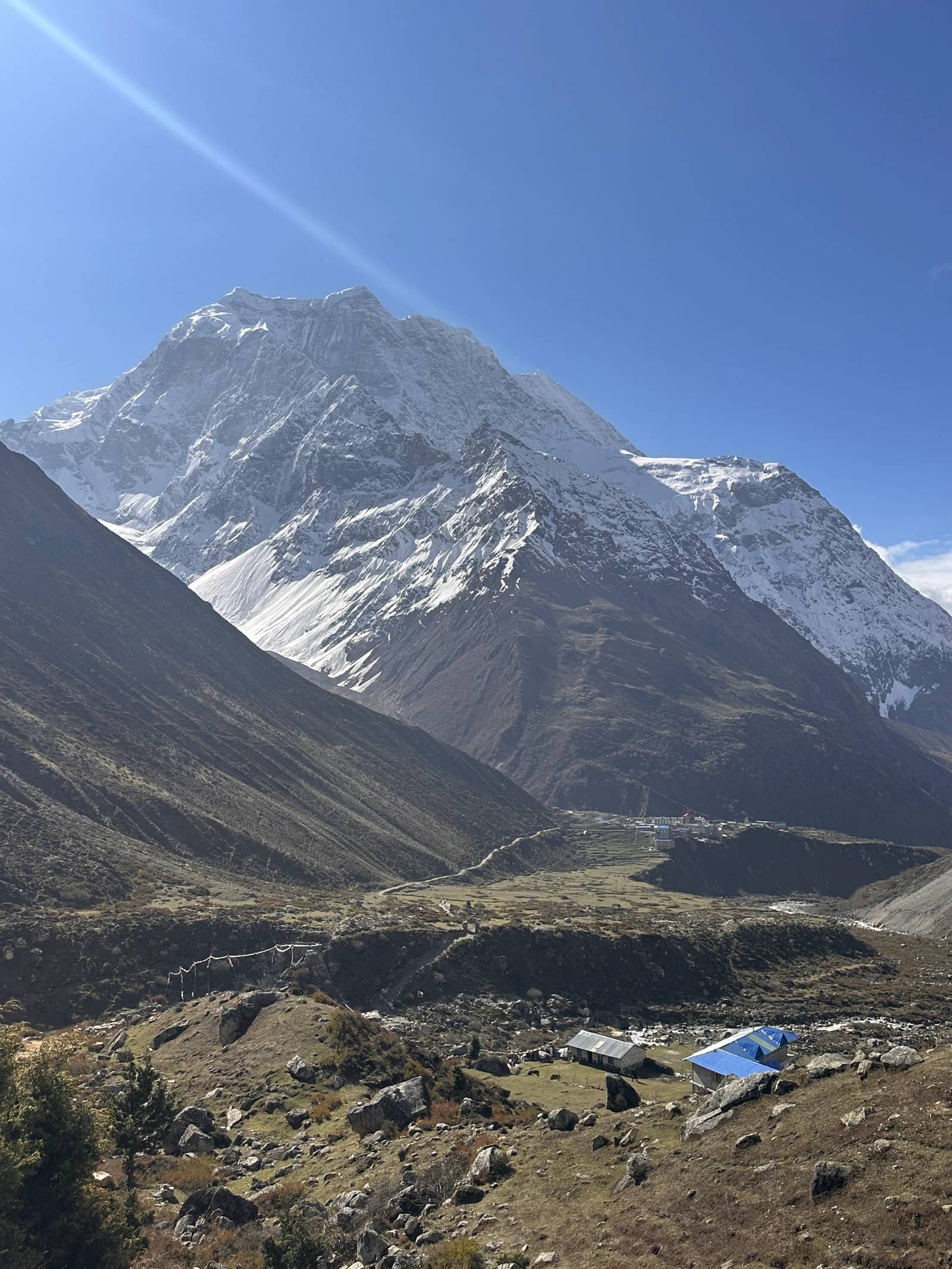 A rugged valley with scattered stone houses leads toward a massive snow-capped mountain under a clear blue sky.